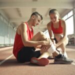 A woman helps an injured man's leg on an indoor track, providing first aid.