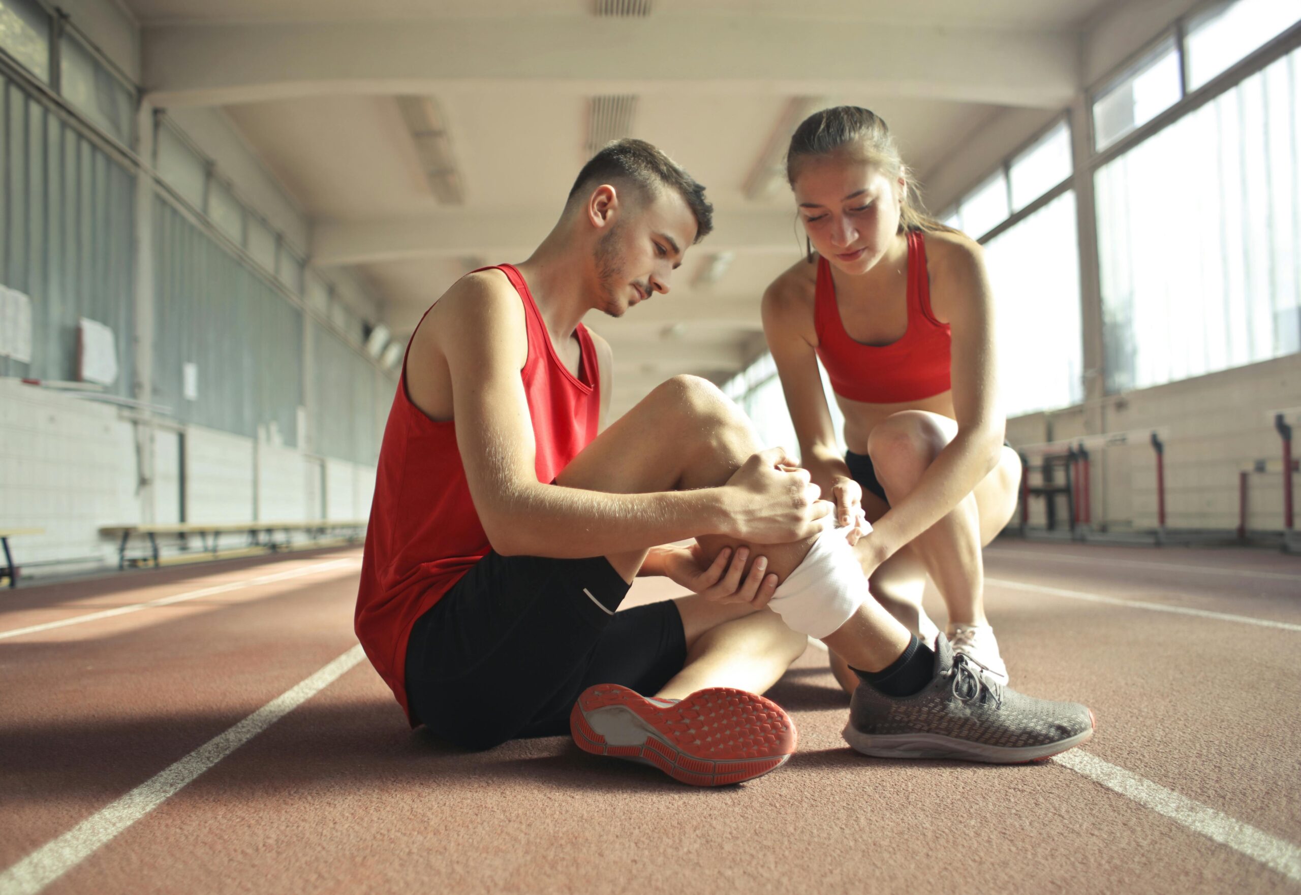 A woman helps an injured man's leg on an indoor track, providing first aid.